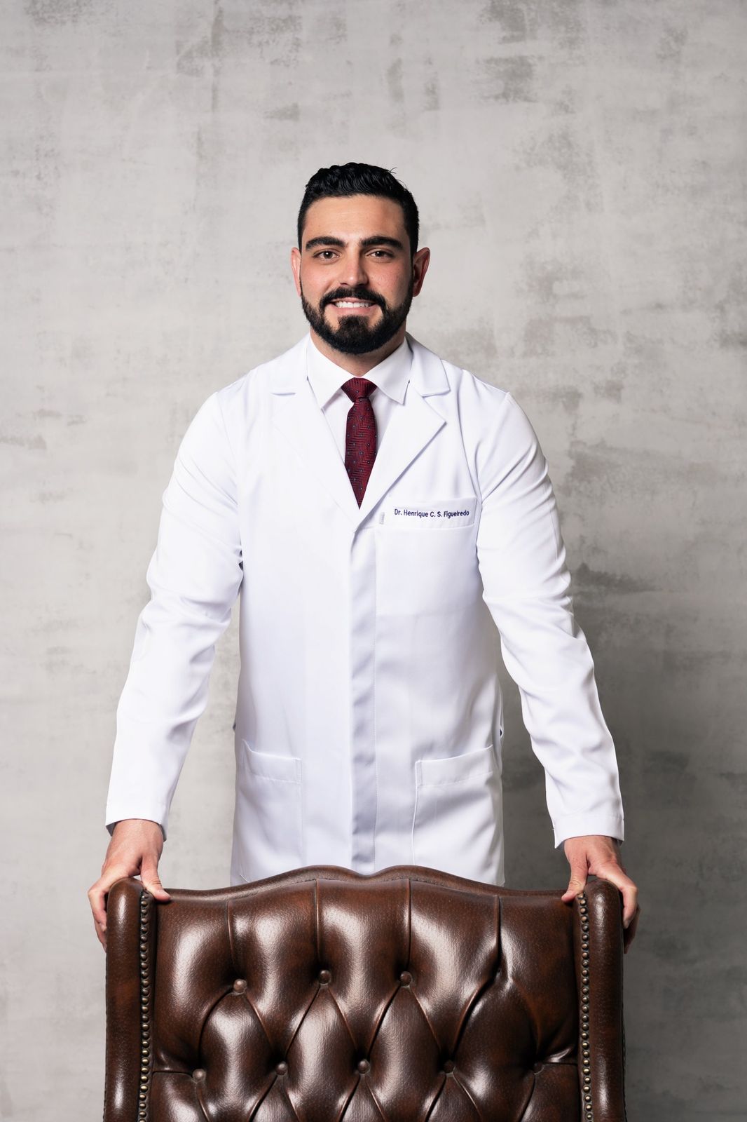Dr. Henrique Figueiredo, de jaleco, apoiado em uma poltrona de couro
